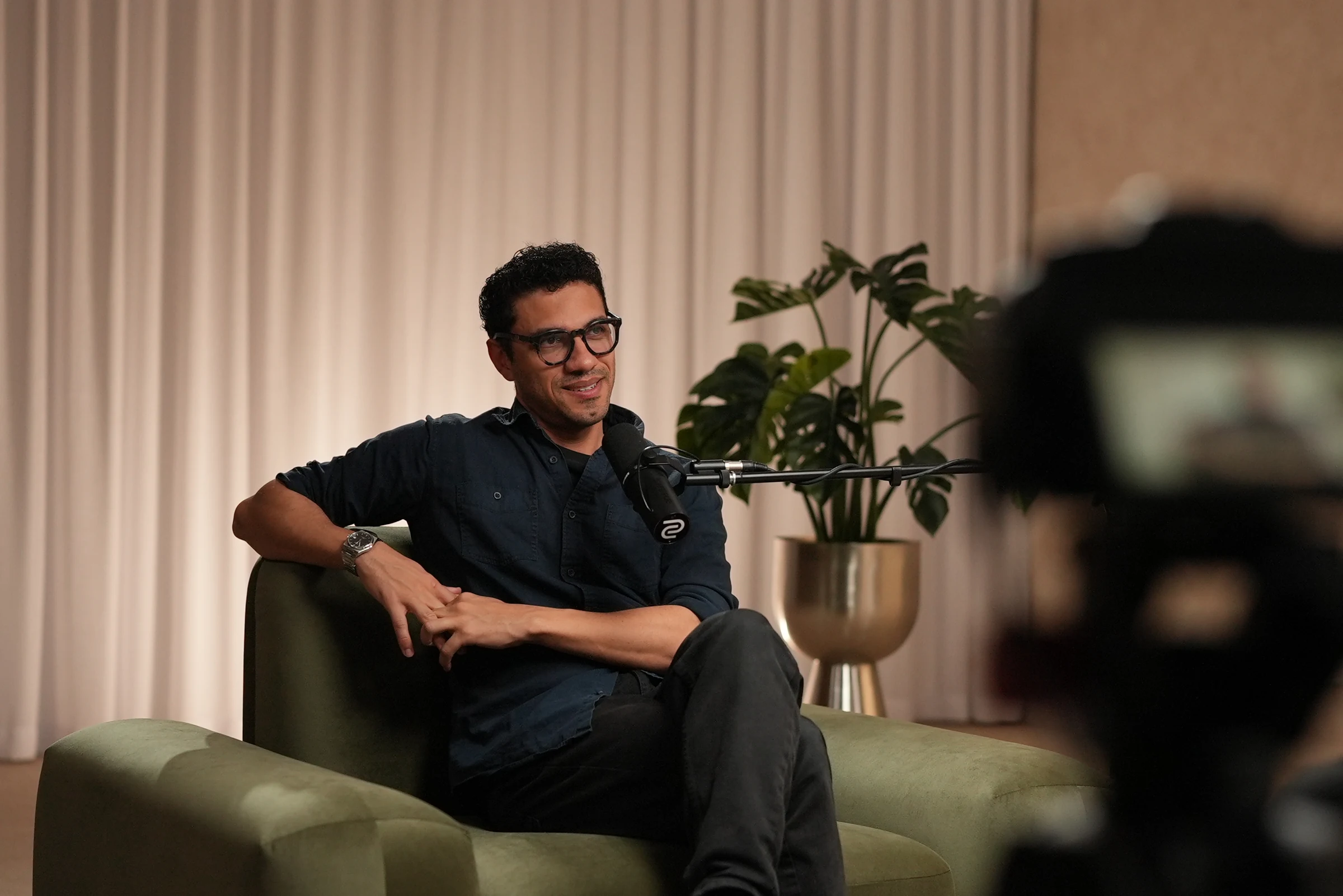 An architect seated in an armchair on set, speaking into a boom microphone, with a camera in the foreground filming the interview.