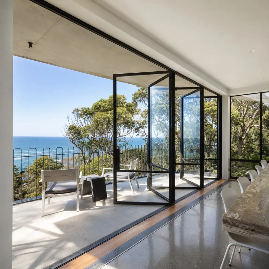 Inside the Guerilla Bay house by DNA Architects, with steel framed glass walls folded open onto a terrace looking across treetops to the ocean.